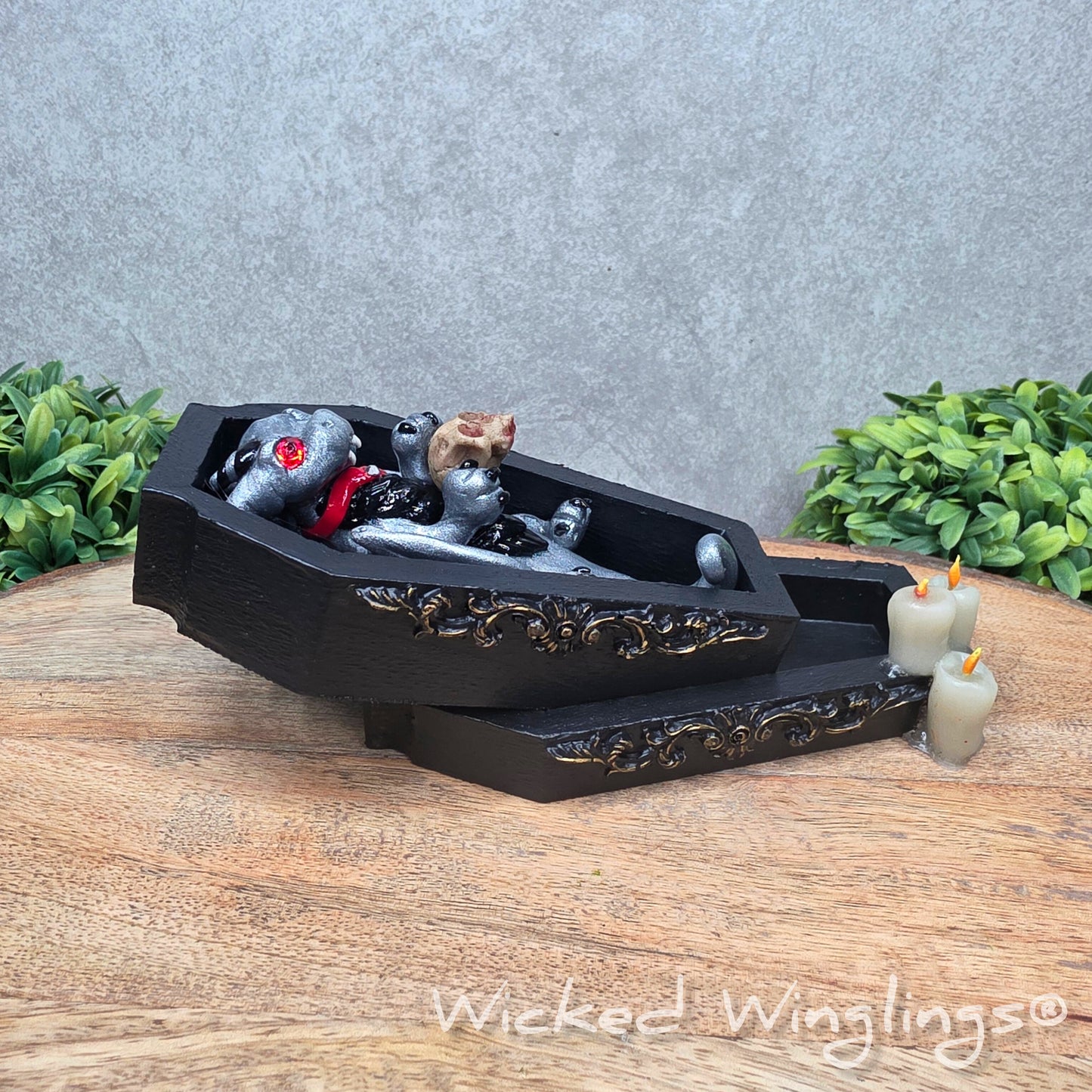 A clay dragon in a black wood coffin with candles and a skull on a wood surface with greenery. Includes text Wicked Winglings