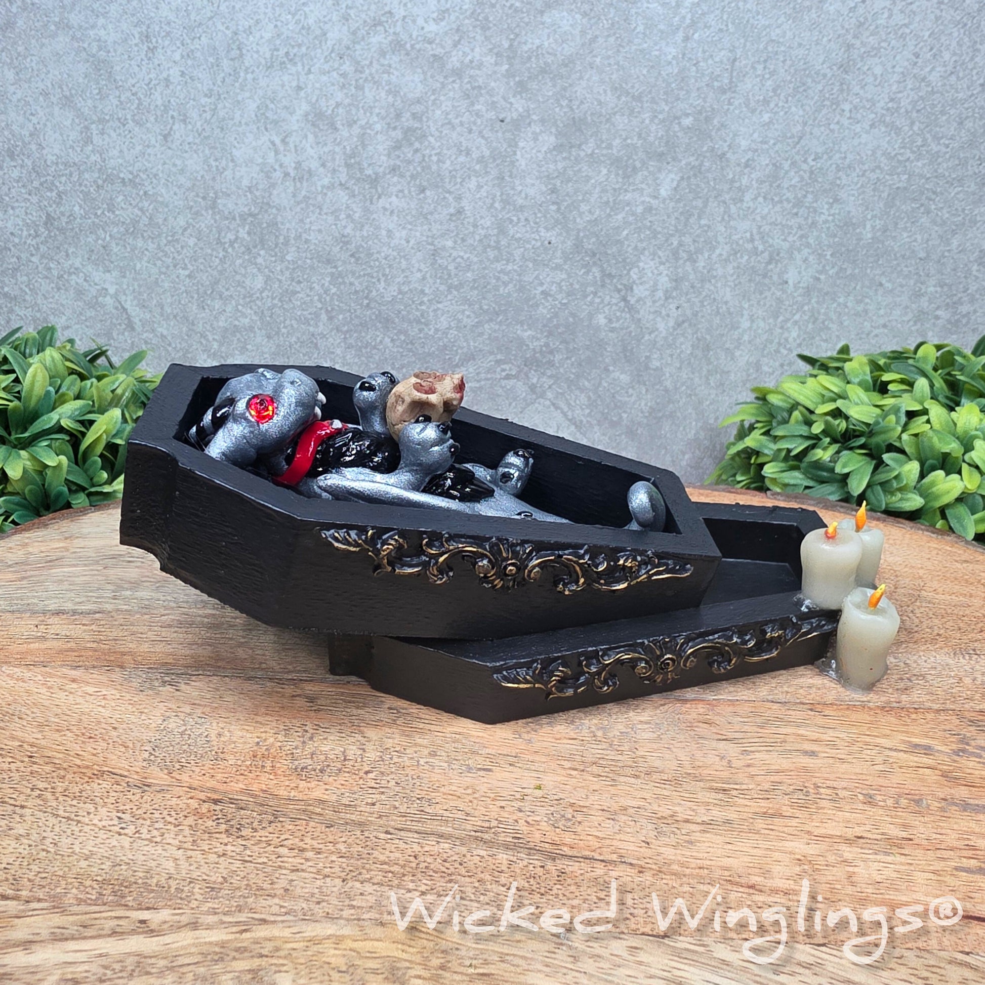 A clay dragon in a black wood coffin with candles and a skull on a wood surface with greenery. Includes text Wicked Winglings
