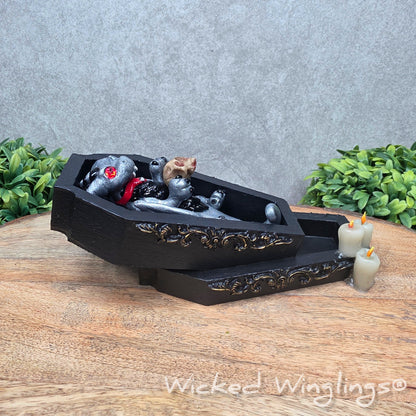 A clay dragon in a black wood coffin with candles and a skull on a wood surface with greenery. Includes text Wicked Winglings