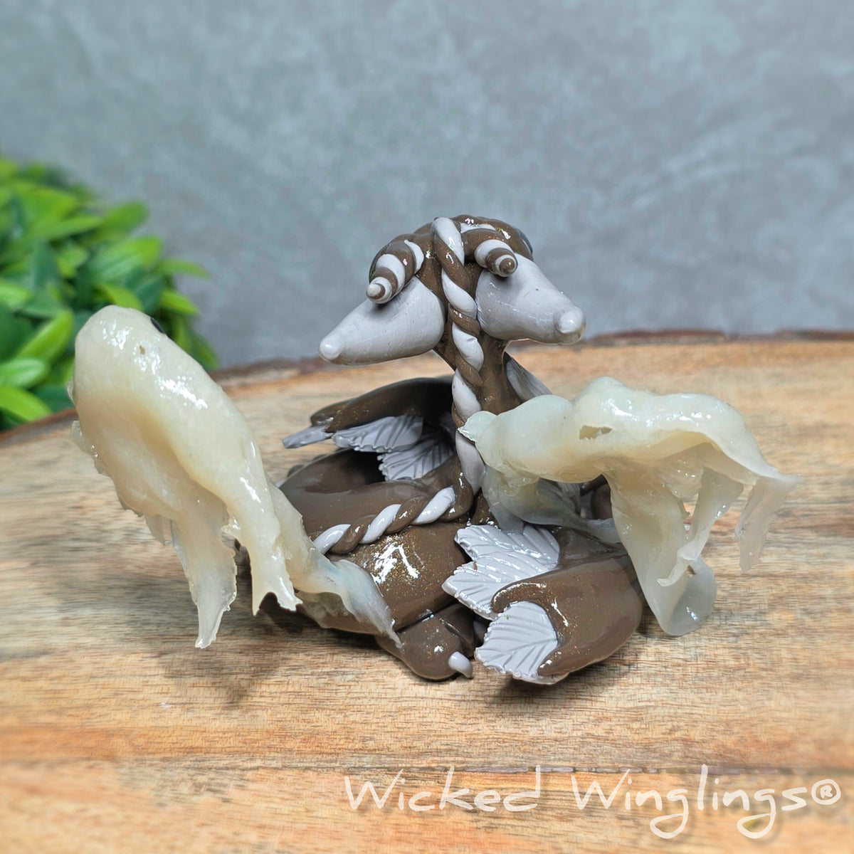 Back view of a  brown clay dragon with ghosts on a wood surface with greenery. Text reads Wicked Winglings.