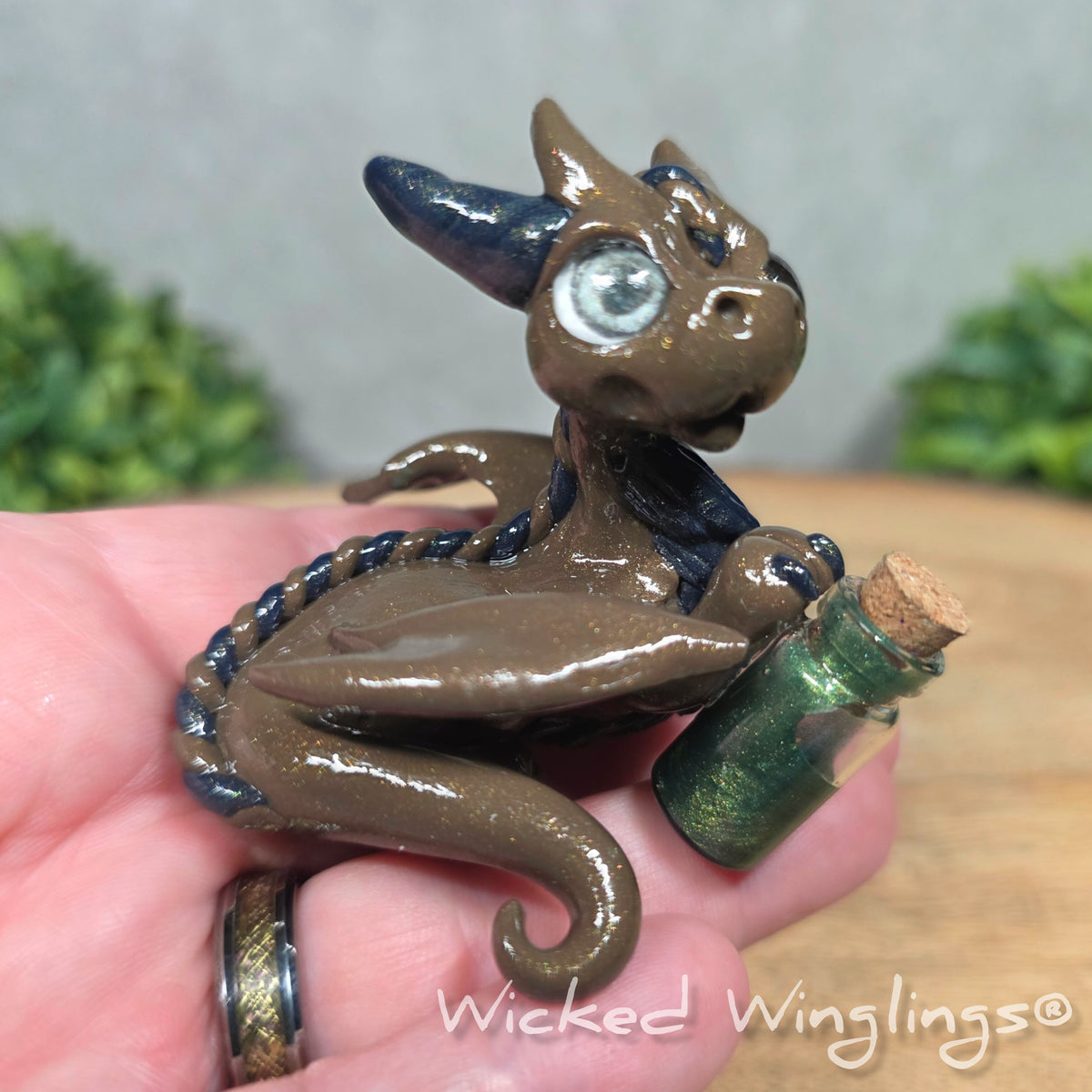 A hand holding a clay dragon with a potion bottle on a wood surface. Text reads Wicked Winglings.