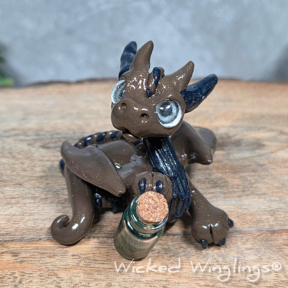 A clay dragon with a potion bottle on a wood surface. Text reads Wicked Winglings.