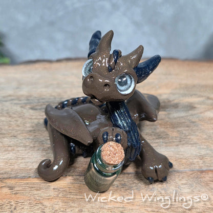 A clay dragon with a potion bottle on a wood surface. Text reads Wicked Winglings.