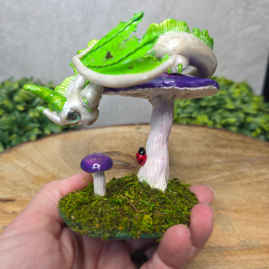 Hand holding  a dragon figurine on a  mushroom with a ladybug on a wooden surface with greenery.