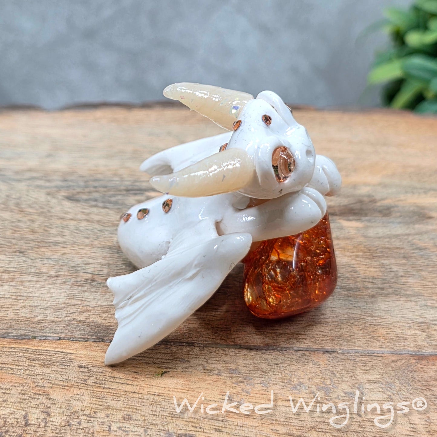 A baby clay dragon sculpture with a crackled quartz against a wood background with greenery and text Wicked Winglings