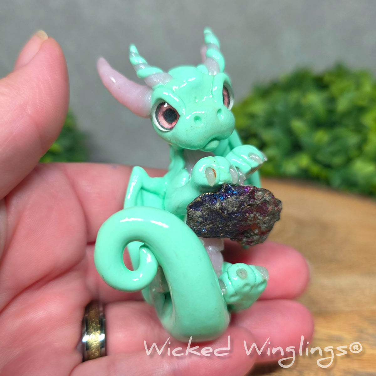 A hand holding a mint clay dragon holding a rainbow colored stone against a blurred background with greenery. Text says Wicked Winglinng.