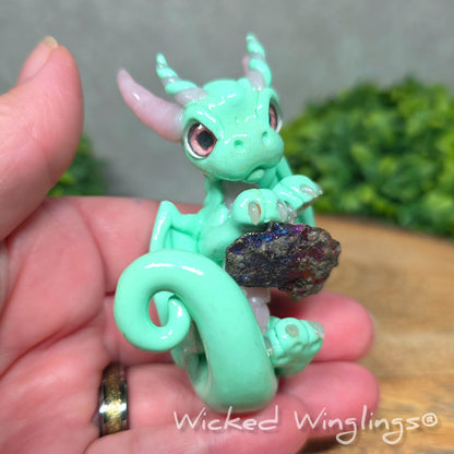 A hand holding a mint clay dragon holding a rainbow colored stone against a blurred background with greenery. Text says Wicked Winglinng.