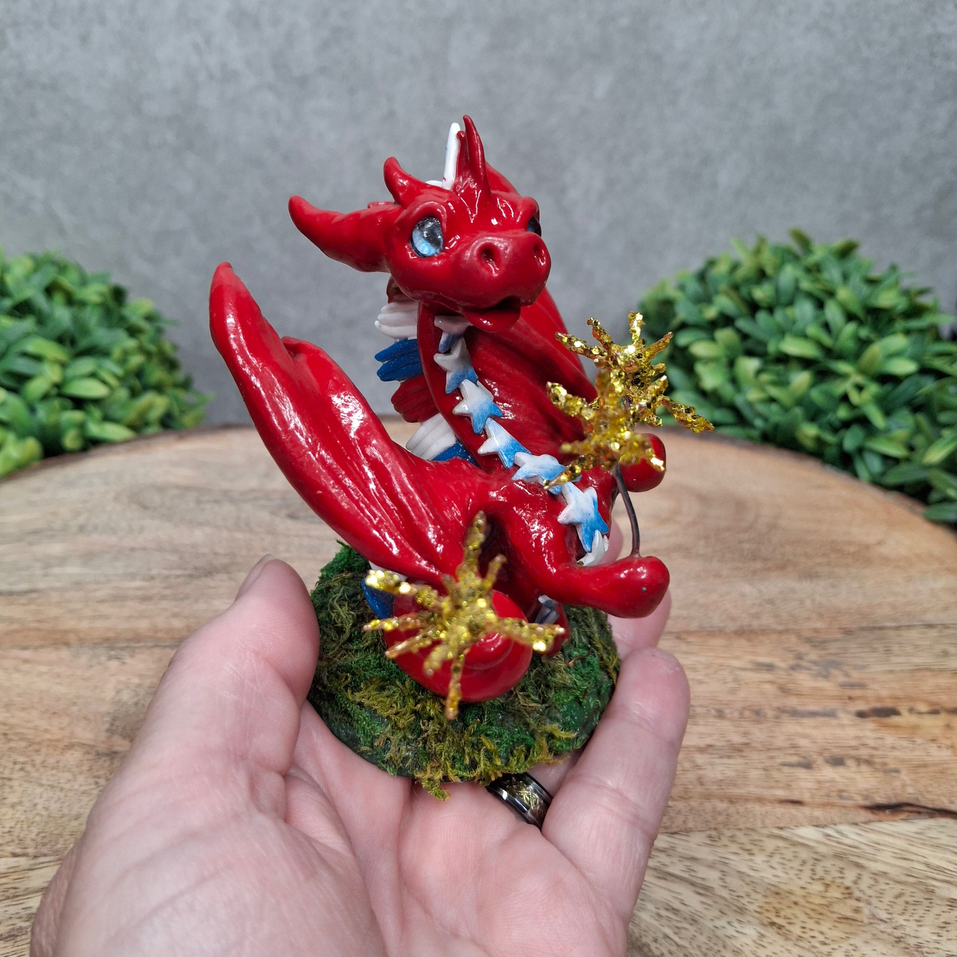 A hand holding a clay dragon with sparklers against a wood surface with greenery
