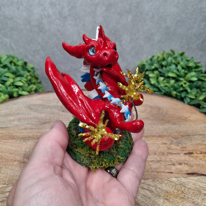 A hand holding a clay dragon with sparklers against a wood surface with greenery