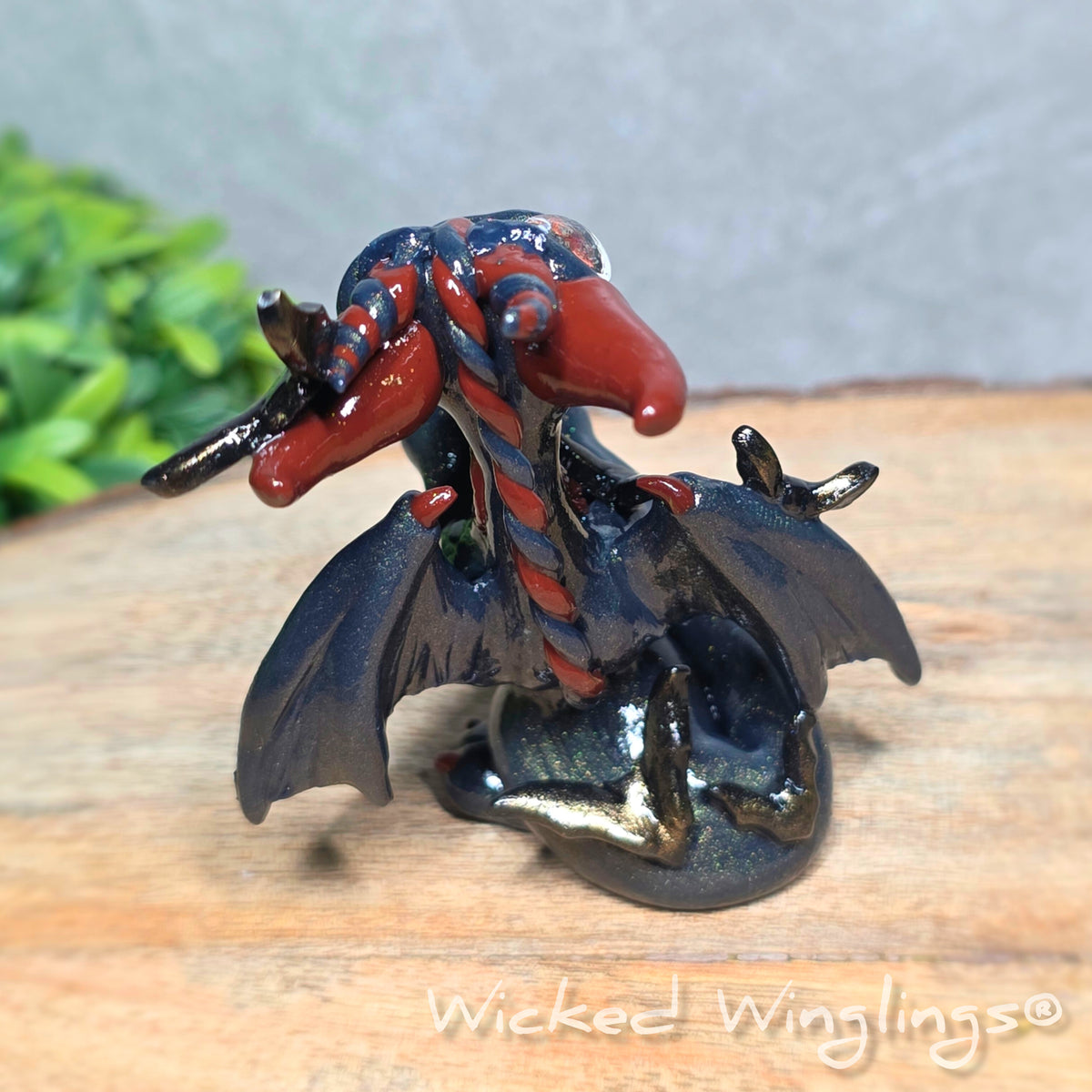 Back view of a clay dragon with bats sitting on a wood surface with greenery. Text reads Wicked Winglings.