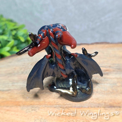 Back view of a clay dragon with bats sitting on a wood surface with greenery. Text reads Wicked Winglings.