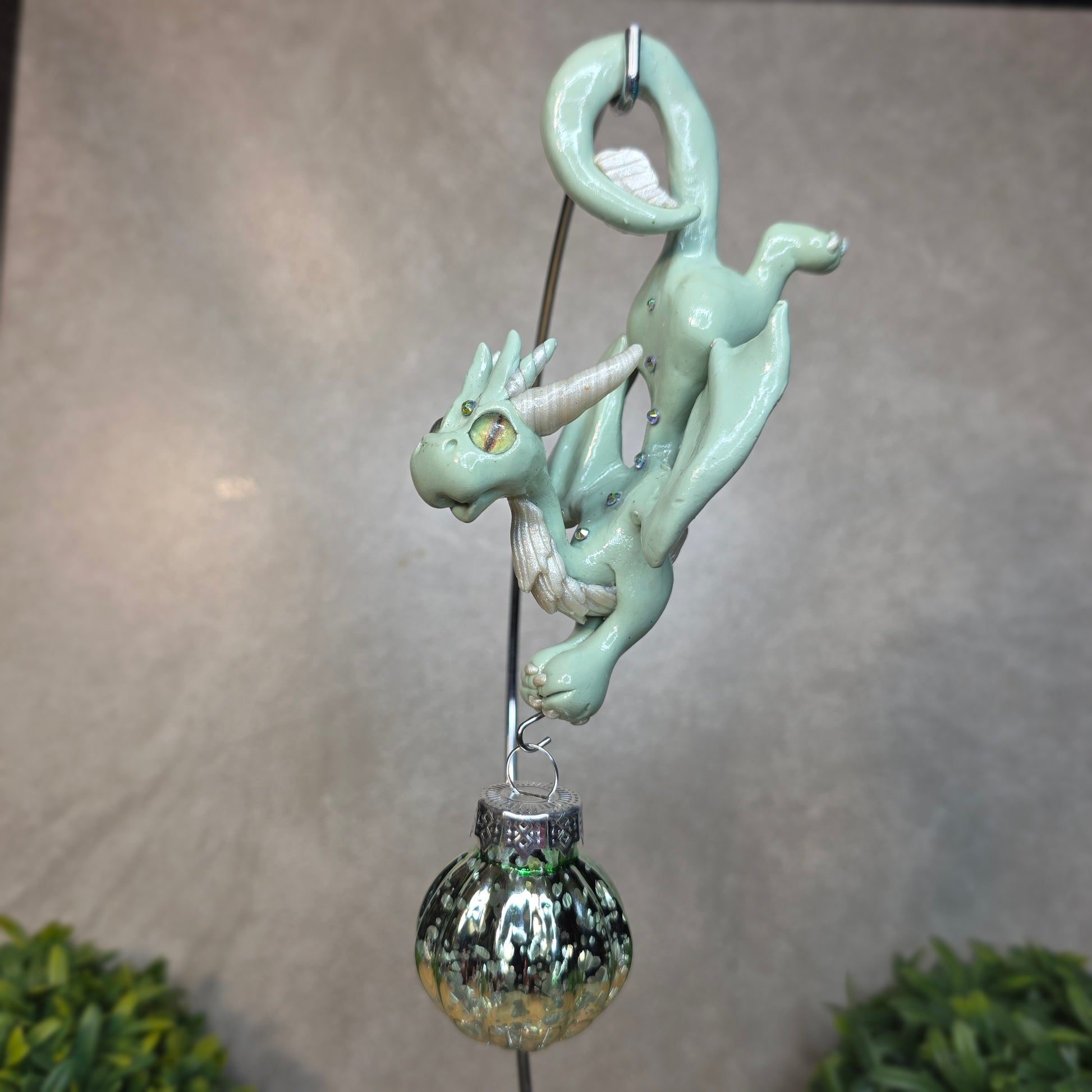 A clay aqua dragon with glass ornament against a blurred background hanging from a stand