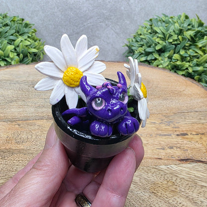 Hand holding a clay baby dragon in a pot with daisies against a wood surface with greenery.