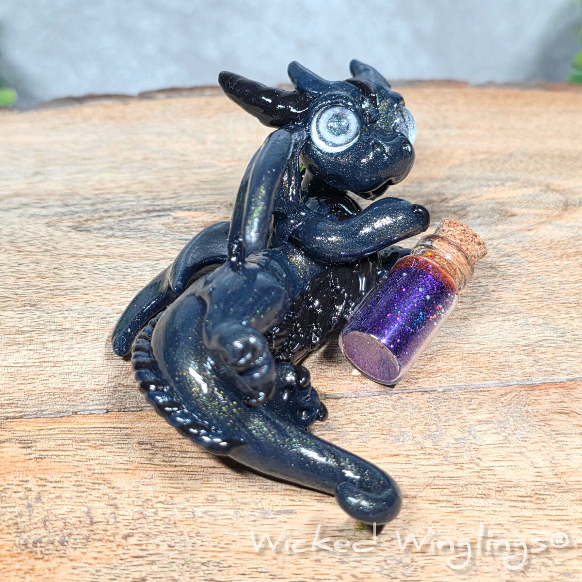 A clay dragon with a potion bottle on a wood surface. Text reads Wicked Winglings.