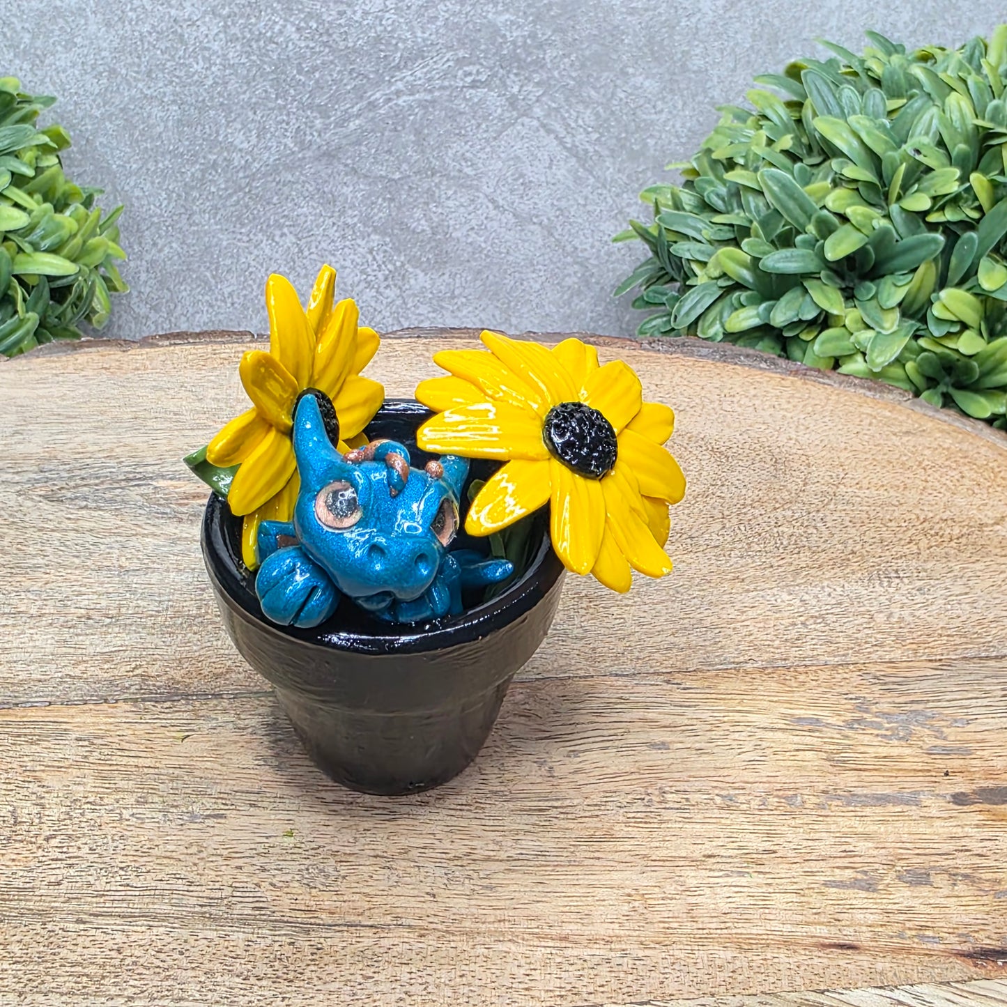 A clay baby dragon in a pot with black eye susans against a wood surface with greenery.
