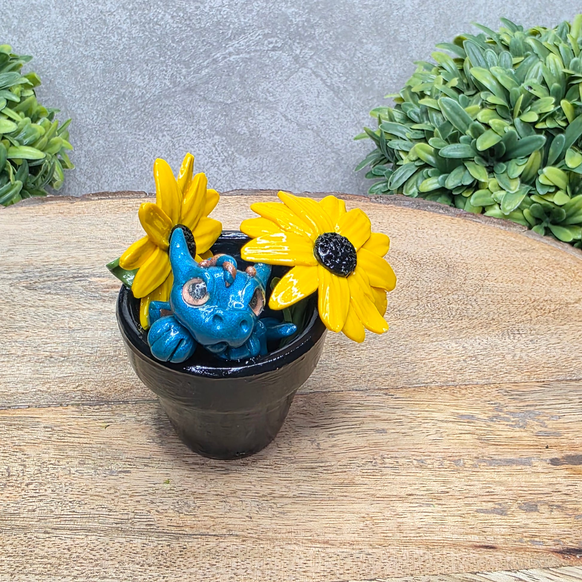 A clay baby dragon in a pot with black eye susans against a wood surface with greenery.