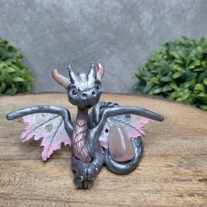 A clay dragon with a rose quartz crystal against a wood surface with greenery.