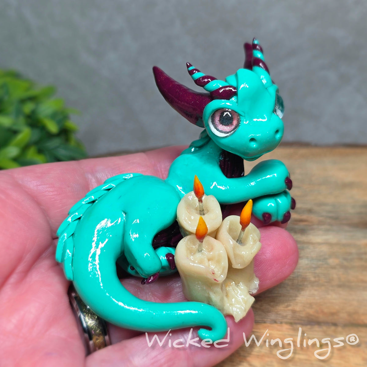 A hand holding a clay dragon with candles against a wood surface with greenery. Text reads Wicked Winglings.