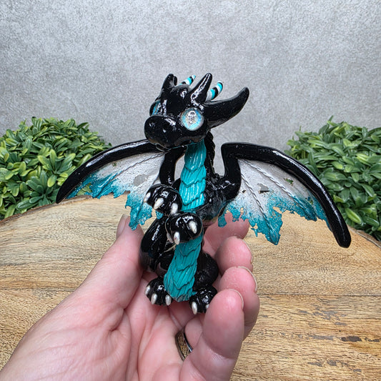 A hand holding a clay dragon against a wood background with greenery