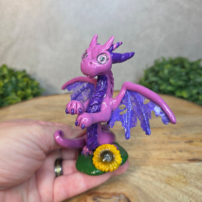 A hand holding clay dragon figurine  with a sunflower on a wooden surface with a blurred green plant in the background