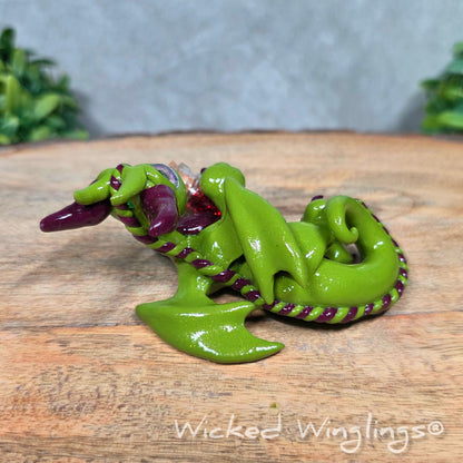 A clay dragon with a potion bottle on a wood surface. Text reads Wicked Winglings.