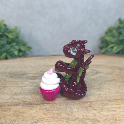 A baby polymer clay dragon with a cupcake against a blurred background