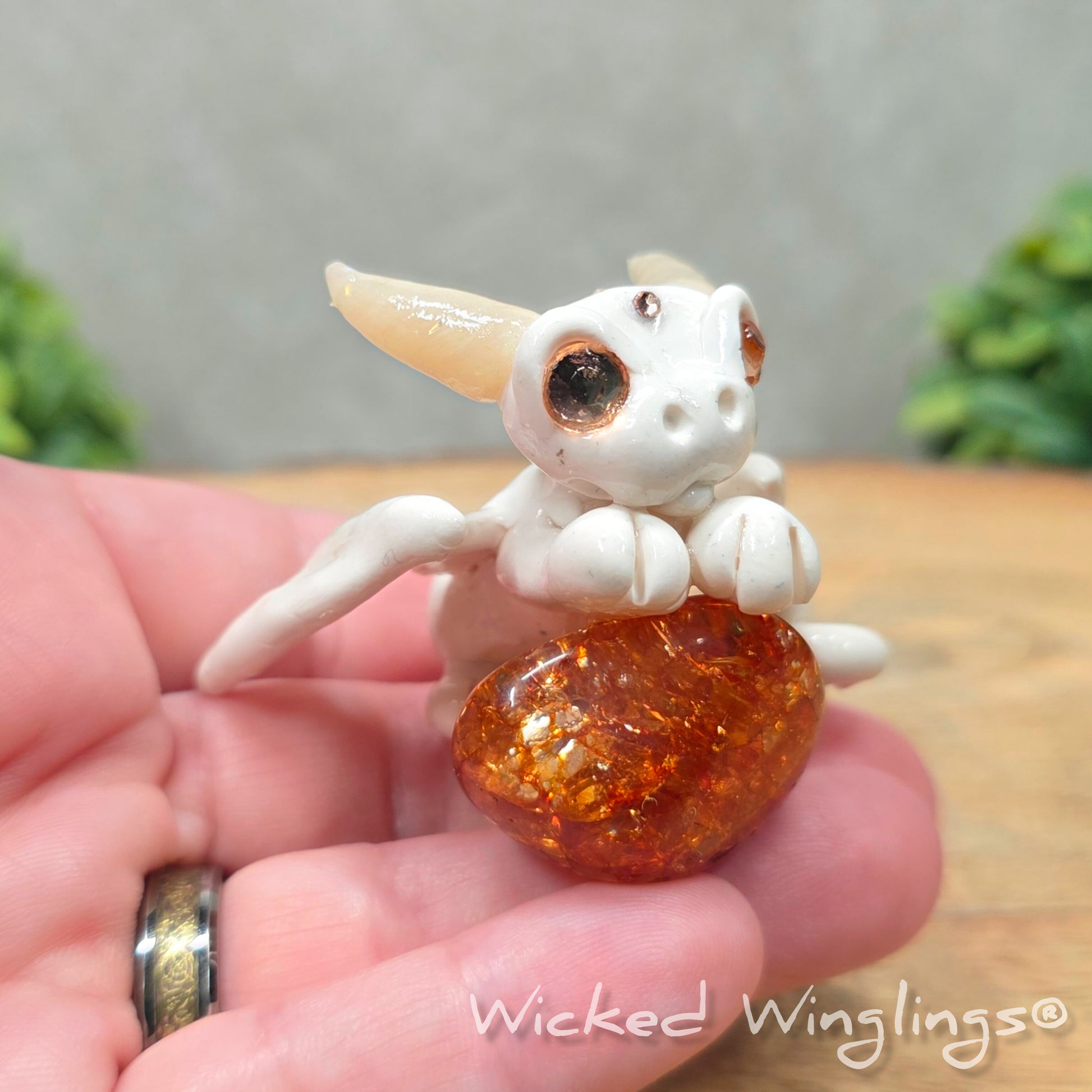 A hand holding a baby clay dragon sculpture with a crackled quartz against a wood background with greenery and text Wicked Winglings