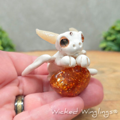 A hand holding a baby clay dragon sculpture with a crackled quartz against a wood background with greenery and text Wicked Winglings