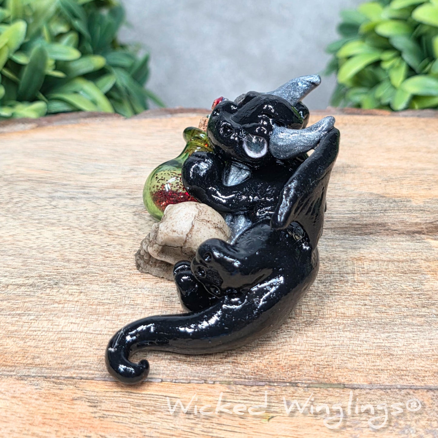 A clay dragon with a skull and potion bottle on a wood surface with greenery. Includes text Wicked Winglings