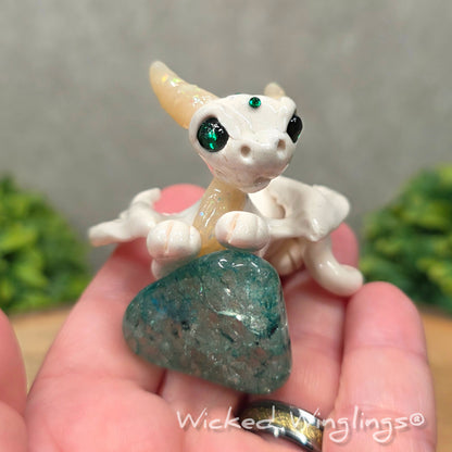 A hand holding a baby clay dragon sculpture with a crackled quartz against a wood background with greenery and text Wicked Winglings