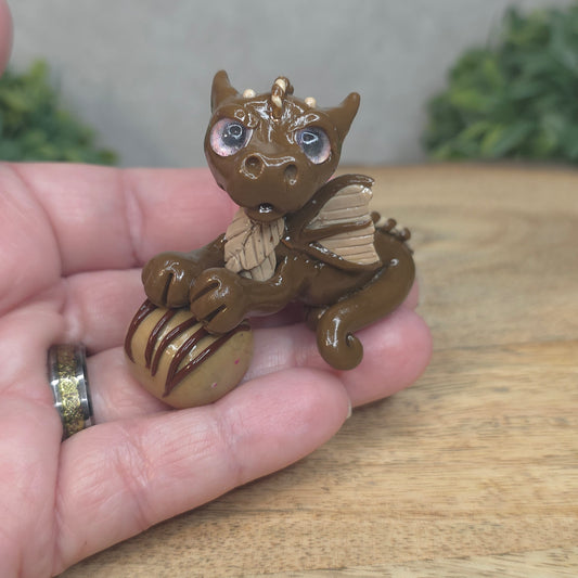 A hand holding a baby polymer clay dragon with a chocolate truffle against a blurred background