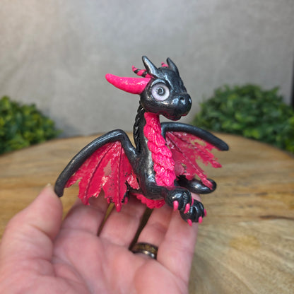A hand holding a graphite and pink clay dragon against a blurred wood surface