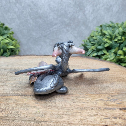 Back view of a clay dragon with a rose quartz crystal against a wood surface with greenery.