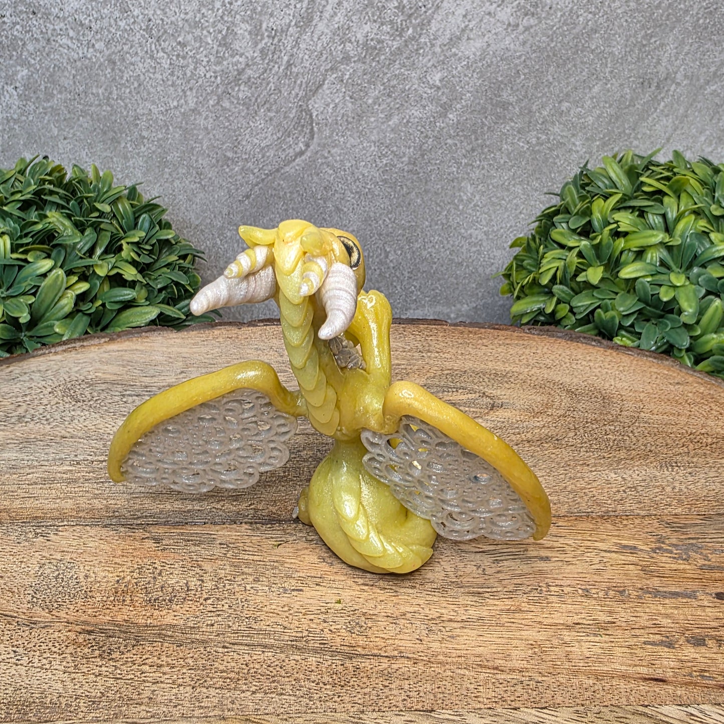 A clay dragon sculpture with lacy wings on a wood surface with greenery.