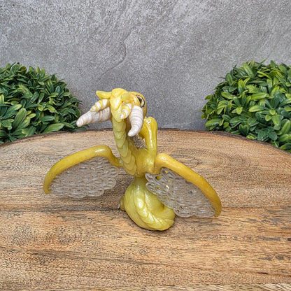 A clay dragon sculpture with lacy wings on a wood surface with greenery.