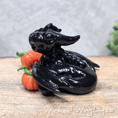 A baby clay dragon sculpture with pumpkins against a wood background with greenery and text Wicked Winglings