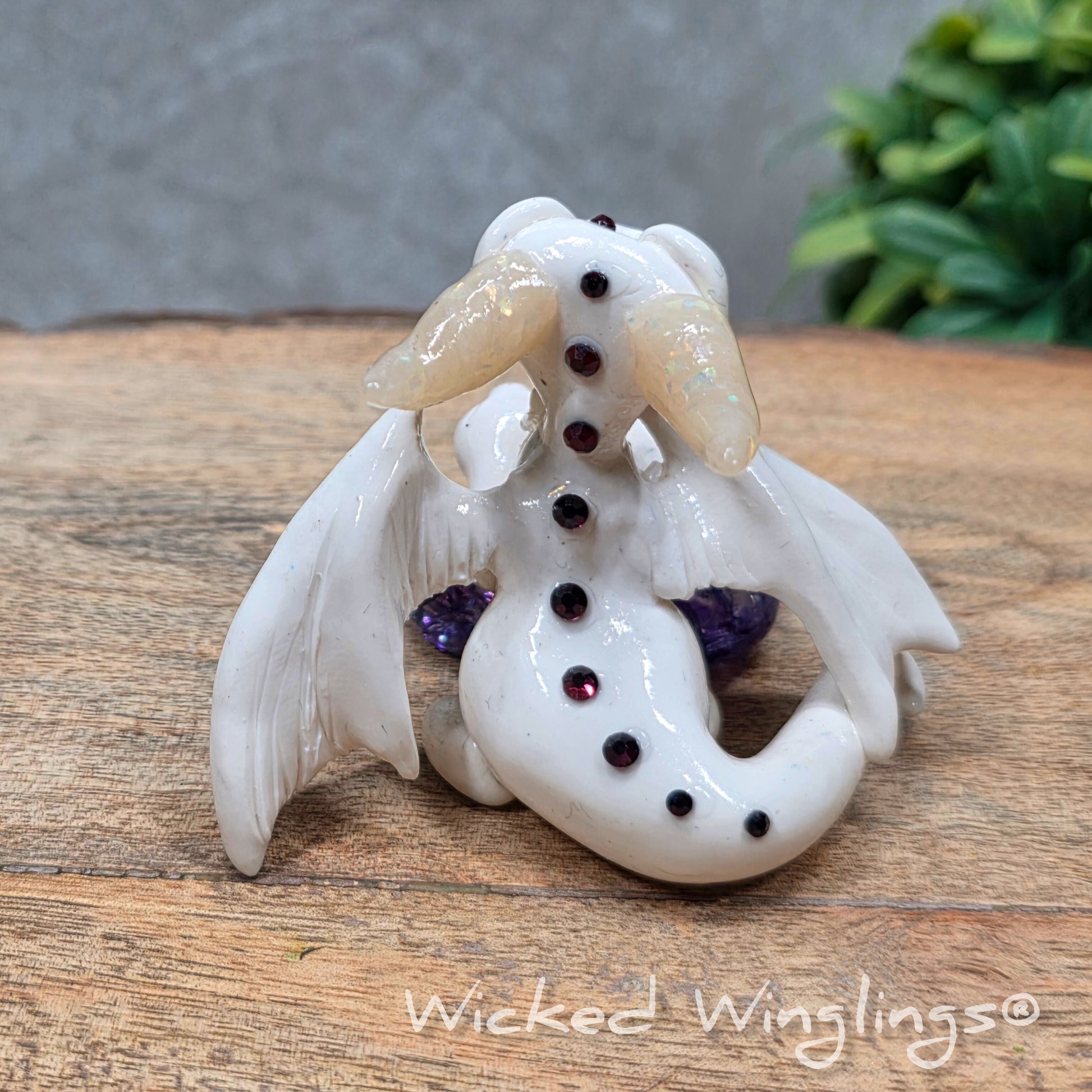 A baby clay dragon sculpture with a crackled quartz against a wood background with greenery and text Wicked Winglings