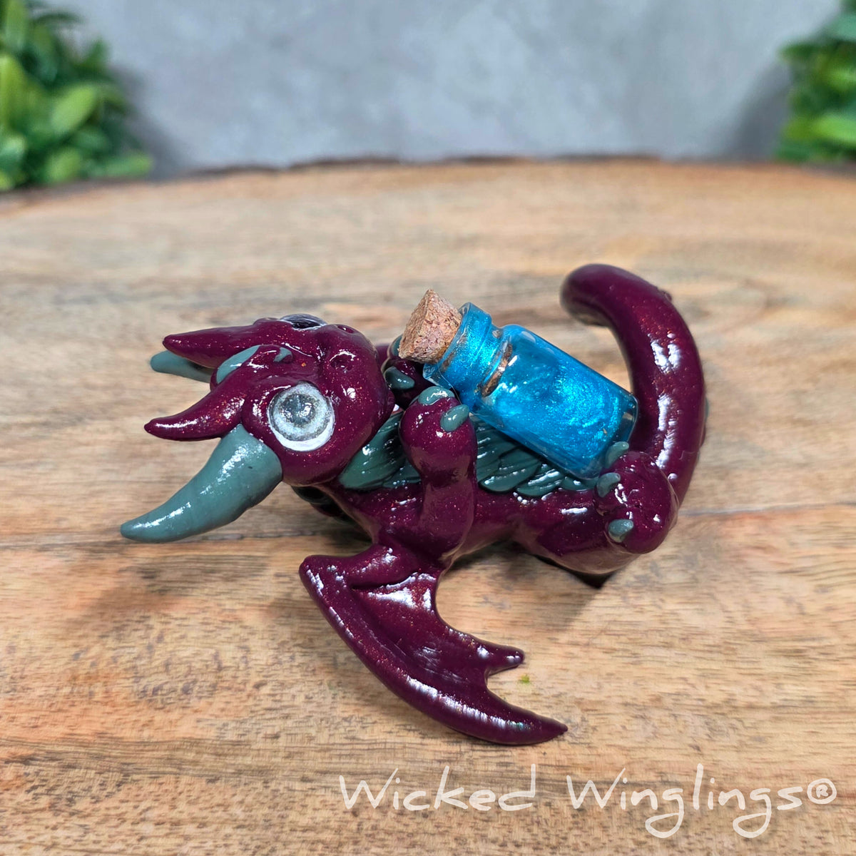 A clay dragon with a potion bottle on a wood surface. Text reads Wicked Winglings.