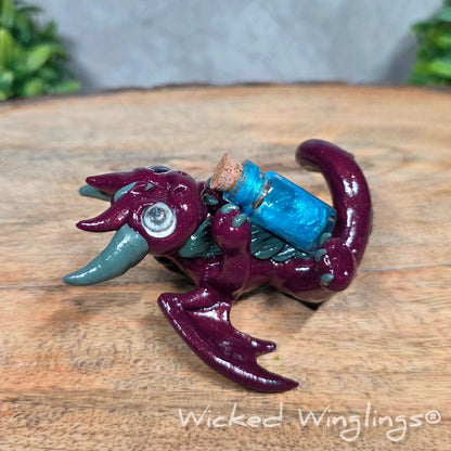 A clay dragon with a potion bottle on a wood surface. Text reads Wicked Winglings.