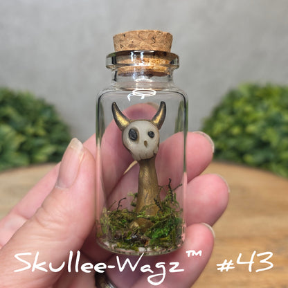 Hand holding a skull faced creature in a jar against a wood background with greenery