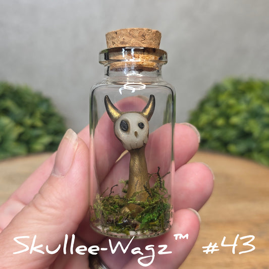 Hand holding a skull faced creature in a jar against a wood background with greenery