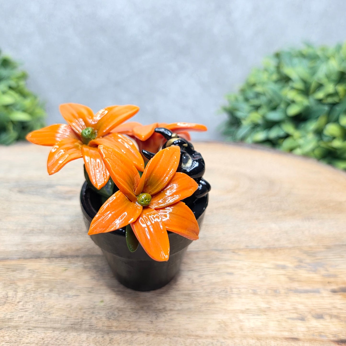 Side view of a clay baby dragon in a pot with lilies against a wood surface with greenery.