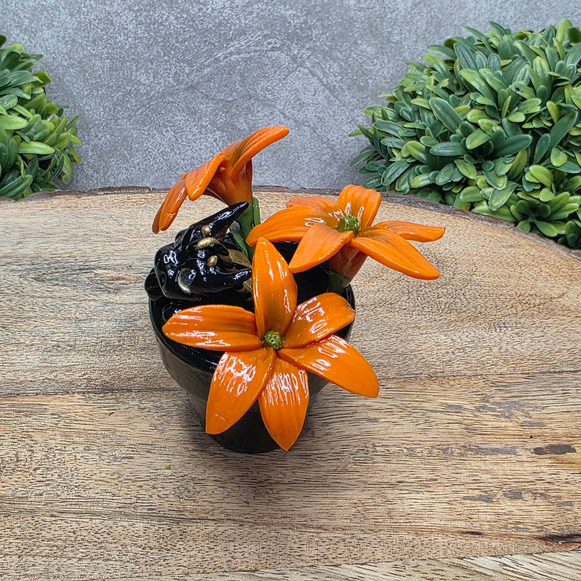 Side view of a clay baby dragon in a pot with lilies against a wood surface with greenery.