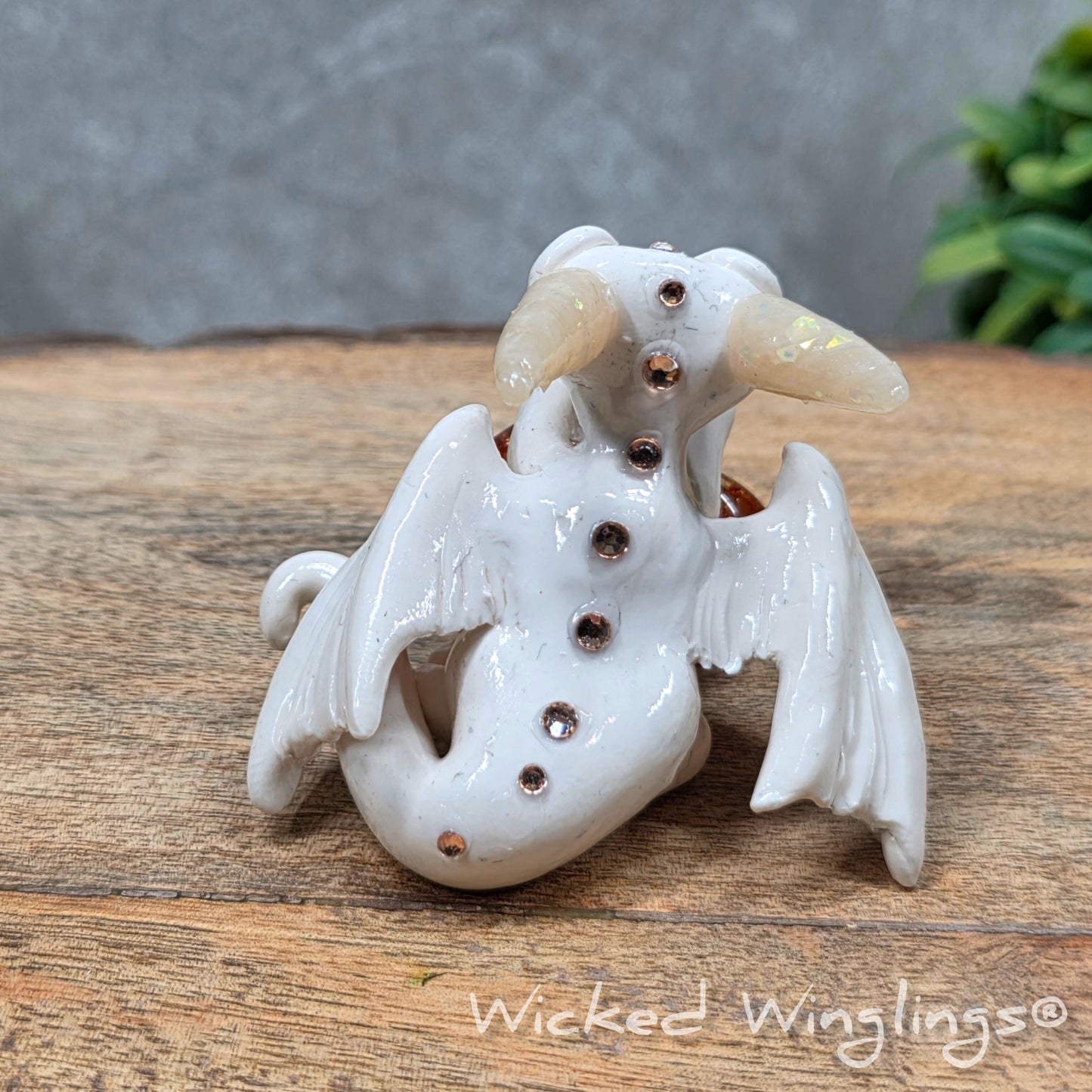 A baby clay dragon sculpture with a crackled quartz against a wood background with greenery and text Wicked Winglings