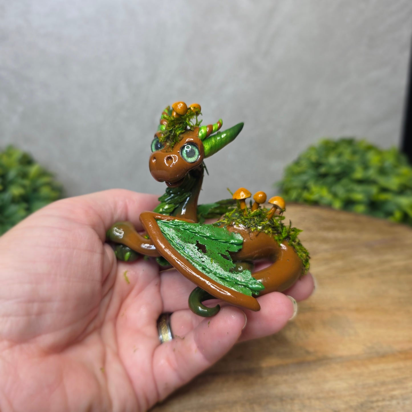 Hand holding a small dragon figurine with green and brown colors against a neutral background