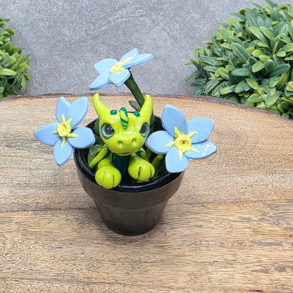 A clay baby dragon in a pot with forget me nots against a wood surface with greenery.