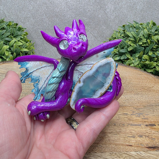 Hand holding a clay dragon with an agate slice against a wood surface with greenery.