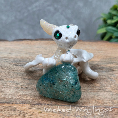 A baby clay dragon sculpture with a crackled quartz against a wood background with greenery and text Wicked Winglings