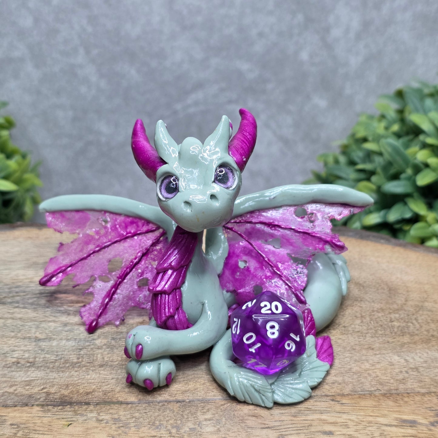 A clay dragon with a D20 dice against a wood surface with greenery.