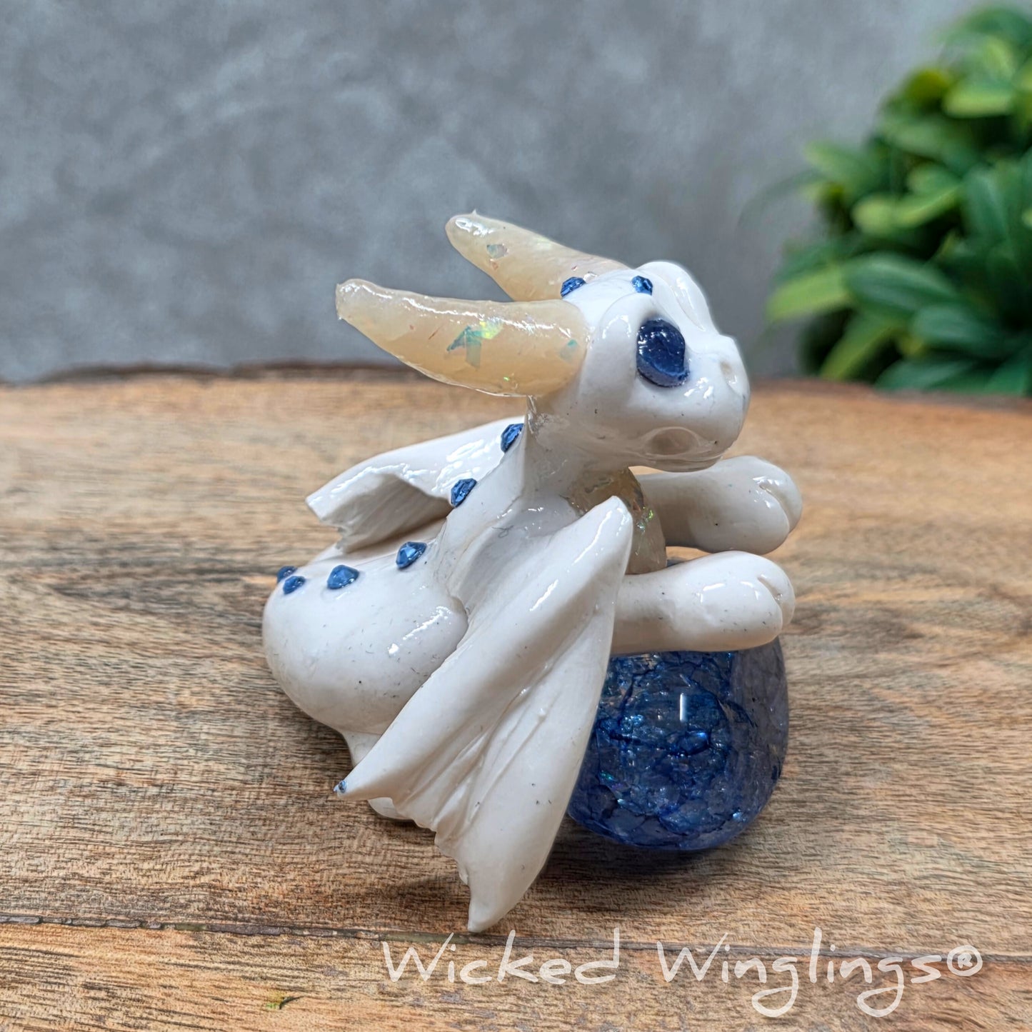 A baby clay dragon sculpture with a crackled quartz against a wood background with greenery and text Wicked Winglings